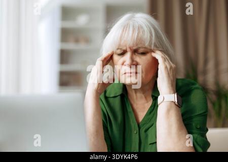 Senior woman suffering from headache while sitting at laptop at home Stock Photo