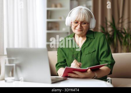 Senior woman writing notes in notebook and using laptop at home Stock Photo