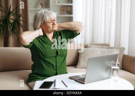Senior woman stretching neck and shoulders after using laptop at home Stock Photo
