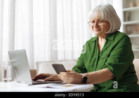 Senior woman using smartphone and laptop at home while checking online information Stock Photo