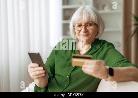 Senior woman sitting on sofa using smartphone and holding credit card Stock Photo
