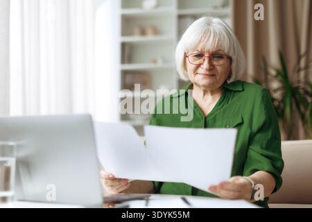 Senior woman reading documents while working on laptop at home Stock Photo
