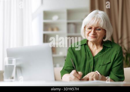 Senior woman writing notes while using laptop at home Stock Photo