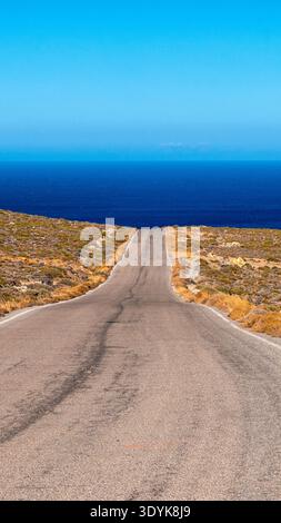 Empty asphalt road leading through dry Mediterranean landscape toward deep blue sea on sunny day. Minimalist travel concept with strong perspective an Stock Photo