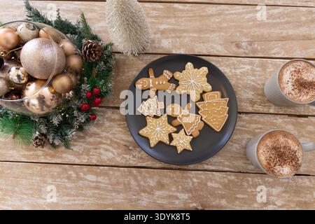 Black plate featuring iced gingerbread cookies on rustic wooden table with wreath and white mugs Stock Photo