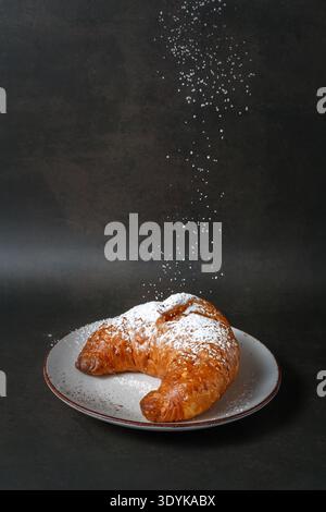 Fresh baked butter croissant on white ceramic plate with falling sugar powder on vintage brown background. Bakery or pastry card with copy space Stock Photo