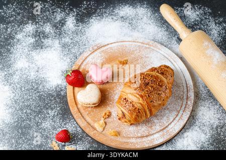 Fresh baked butter crushed nuts breakfast croissant, french heart macarons, strawberry and crumbs  on wooden board over dark table with flour. Kitchen Stock Photo