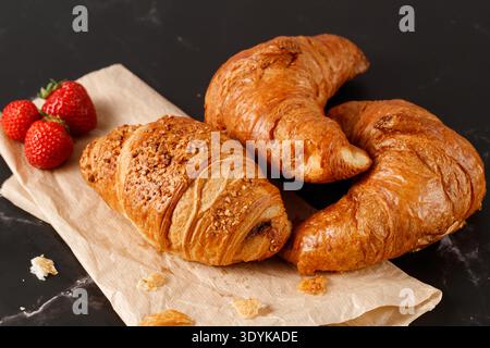 Freshly baked  for breakfast nougat crushed nuts and butter croissants with crumbs and strawberries on paper on a dark marble table Stock Photo