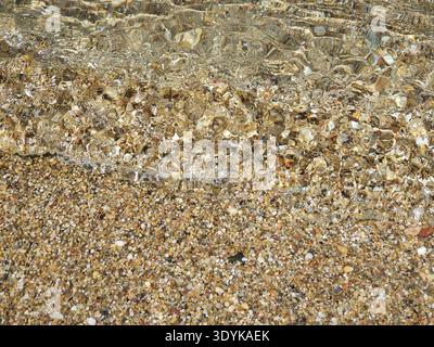 Crystal-clear shallow water gently rippling over a pebble beach, sunlit stones visible beneath the surface creating textured patterns and a calm, peac Stock Photo