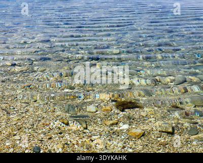 Crystal-clear shallow water gently rippling over a pebble beach, sunlit stones visible beneath the surface creating textured patterns and a calm, peac Stock Photo