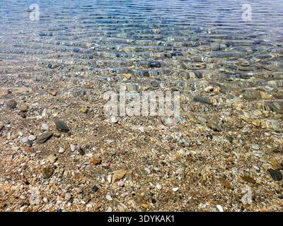 Crystal-clear shallow water gently rippling over a pebble beach, sunlit stones visible beneath the surface creating textured patterns and a calm, peac Stock Photo