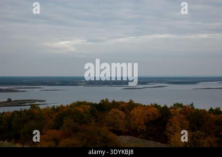 Autumn landscape with dense forest seen from above in green, brown and orange tone. Clouded sky overhead, distant river fragment visible. Moody and ca Stock Photo