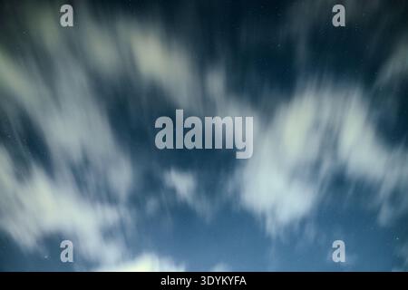 Long exposure capture of night clouds slowly flowing across a deep blue sky, revealing scattered stars between the soft white cloud streaks and creati Stock Photo