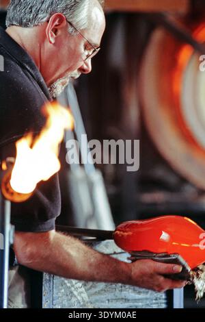 A skilled glassblower works carefully, using tools to shape a glowing red glass piece. The workshop is filled with equipment, and flames rise nearby a Stock Photo