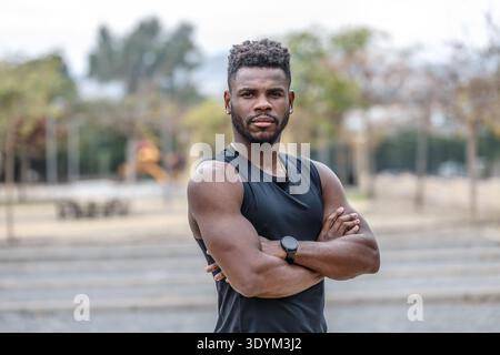 Muscular young black bearded male athlete with wristwatch standing on city street and crossing hands during fitness training Stock Photo
