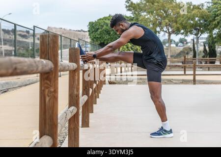Full body side view of strong African American athlete putting leg on wooden fence and doing toe reach exercise during fitness workout Stock Photo