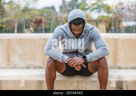 Depressed African American male athlete with hood on head sitting on stairs during break from fitness training in city park Stock Photo