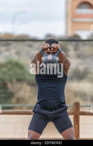 Young strong African American athlete in sports clothes lifting heavy weight while training in city park doing kettlebell swing exercise during daytim Stock Photo