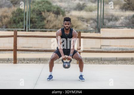Full body of determined African American male athlete doing squats with heavy kettlebell during intense training on sports ground Stock Photo