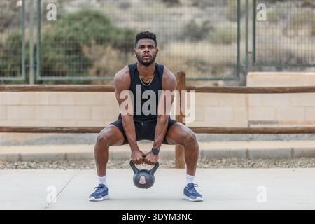 Full body of determined African American male athlete in activewear doing squats with heavy kettlebell during intense weightlifting training Stock Photo