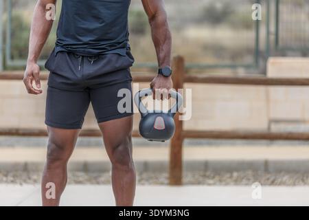 Crop anonymous African American athlete in activewear standing against fence on sports ground and holding heavy kettlebell during functional training Stock Photo