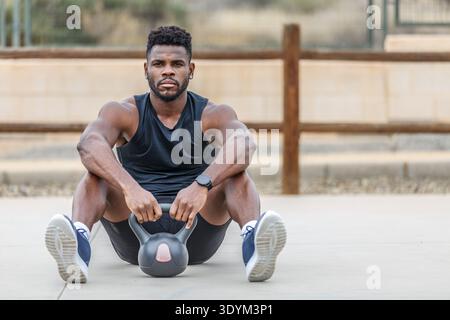 Bearded sportsman in sneakers and activewear with kettlebell sitting on sports ground against blurred background while looking at camera during fitnes Stock Photo