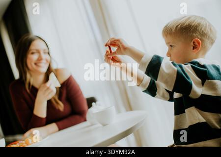 A child sits at a table in a cafe and holds up a snack while his mother smiles nearby. They seem to be having a nice time sharing food and enjoying ea Stock Photo