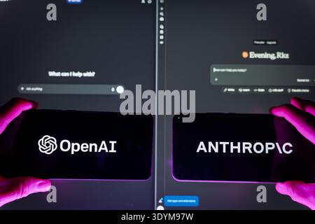 Person holding two smartphones displaying OpenAI and Anthropic logos against computer screens with AI chat interfaces, artificial intelligence competi Stock Photo