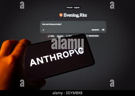 A hand holding a smartphone displaying the Anthropic logo in front of a computer screen showing the Claude AI interface, representing artificial intel Stock Photo