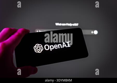 A hand holding a smartphone displaying the OpenAI logo in front of a computer screen showing the ChatGPT interface, representing generative artificial Stock Photo