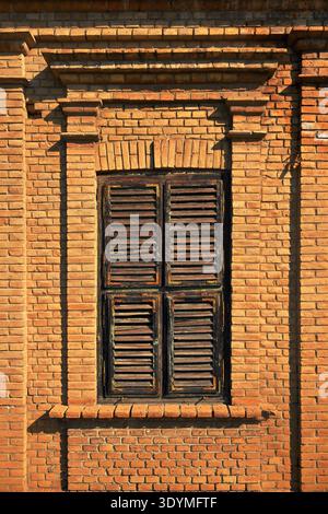 Classic rectangular window set in a rich red brick wall, featuring thick decorative brick lintel, with dark weathered wooden shutters, vertical image. Stock Photo