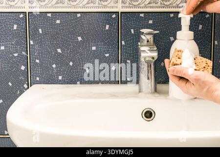 Hand holding foamy sponge while pressing white soap dispenser pump, next to running chrome faucet on white sink with blue mosaic backdrop. Stock Photo
