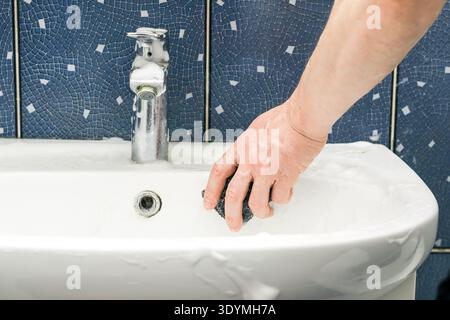 Focus on hand applying soapy sponge to scrub interior of white sink in blue-tiled bathroom Stock Photo