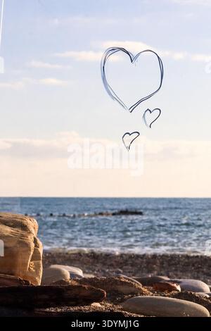 Blue line drawings of hearts arranged like thought bubbles against a tranquil pebble beach and expansive ocean backdrop. Stock Photo