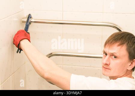 Young man with serious expression in red glove using wrench to adjust chrome rail on white tiled bathroom wall. Stock Photo