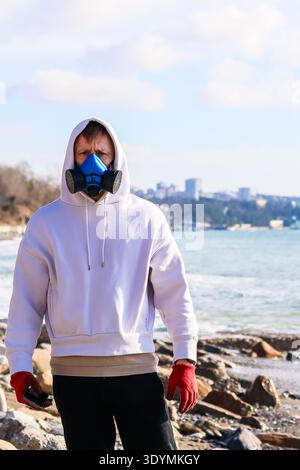 Person in hooded sweatshirt, protective respirator and red gloves standing on pebble beach overlooking calm sea. Stock Photo