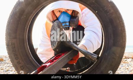 Equipped volunteer uses crowbar to remove debris from old tire on pebble-filled coastline. Stock Photo