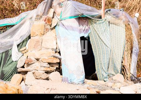 Hand-built hut using rocks, plastic wraps, and assorted textiles as walls and roof, situated among boulders and sparse vegetation on uneven ground. Stock Photo