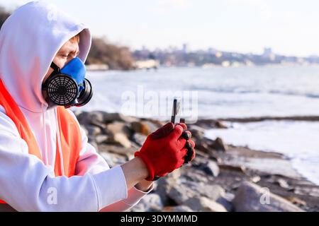 Side profile of worker in safety attire with gas mask using smartphone near waves and distant buildings. Stock Photo