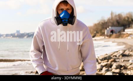 Person wearing protective equipment stands facing camera on stony beach with sea and urban horizon Stock Photo
