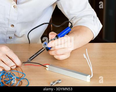 Hands using soldering iron to join red and black wires to silver rectangular module near coiled cables Stock Photo