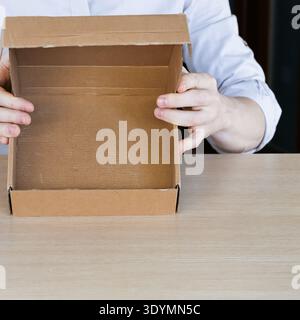Arms raising small open cardboard box above table, displaying empty interior space. Stock Photo