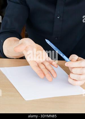 Male hands over wooden table, right fist holding blue pen upward, left palm facing up near white blank paper sheet Stock Photo