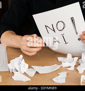 Close-up of hands displaying handwritten protest sign against artificial intelligence amid discarded paper waste on wooden table Stock Photo