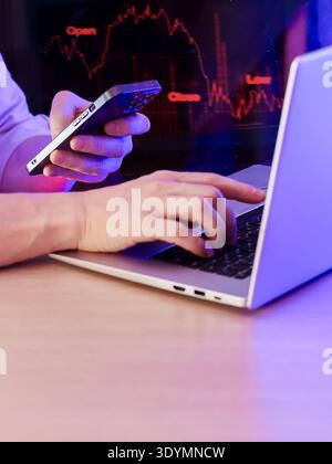 Person typing on laptop while holding smartphone showing red downward graph, red neon financial data in background. Stock Photo