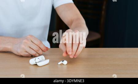 Close-up of hands placing one white wireless earbud beside an open charging case on a light wooden surface in indoor setting Stock Photo
