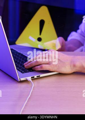 Hands on laptop keyboard in front of blurred yellow triangular warning sign with exclamation mark Stock Photo
