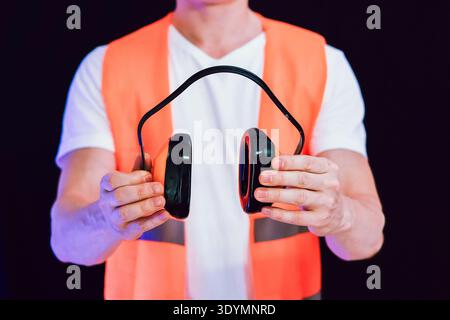 Close-up of hands in orange vest showing black and orange protective earmuffs against black backdrop Stock Photo