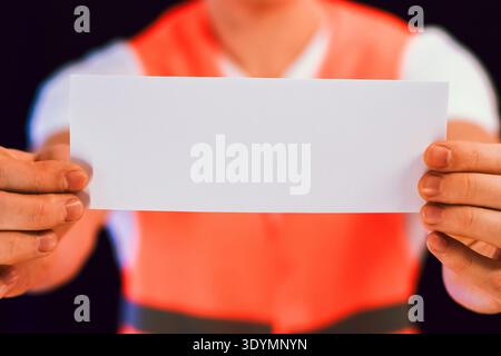 Extreme close view of person's arms in orange reflective vest extending wide blank white rectangular sheet towards viewer on black Stock Photo