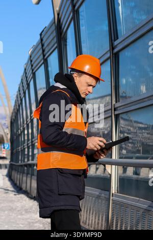 Male engineer in hard hat and vest reviews clipboard while leaning against railing near glass panels reflecting city in winter. Stock Photo
